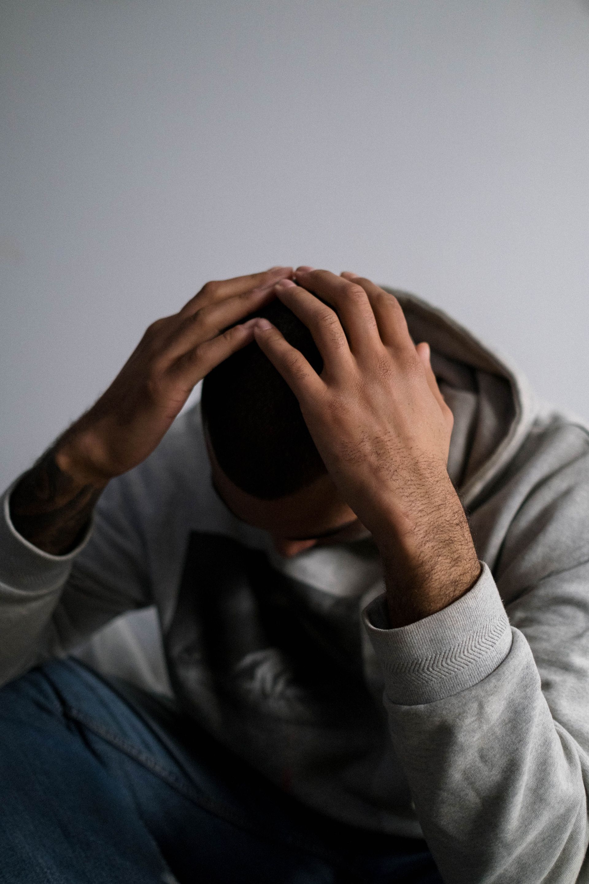 A man in a hoodie sits indoors, appearing contemplative or sad, with a focus on his hands and posture.